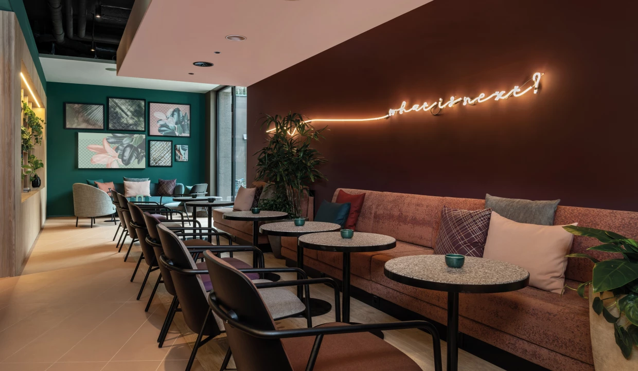 Modernes Hotel-Bistro mit terracottafarbenen Sofas, gepolsterten Stühlen, runden Tischen, Leuchtschriftwand und Pflanzen.