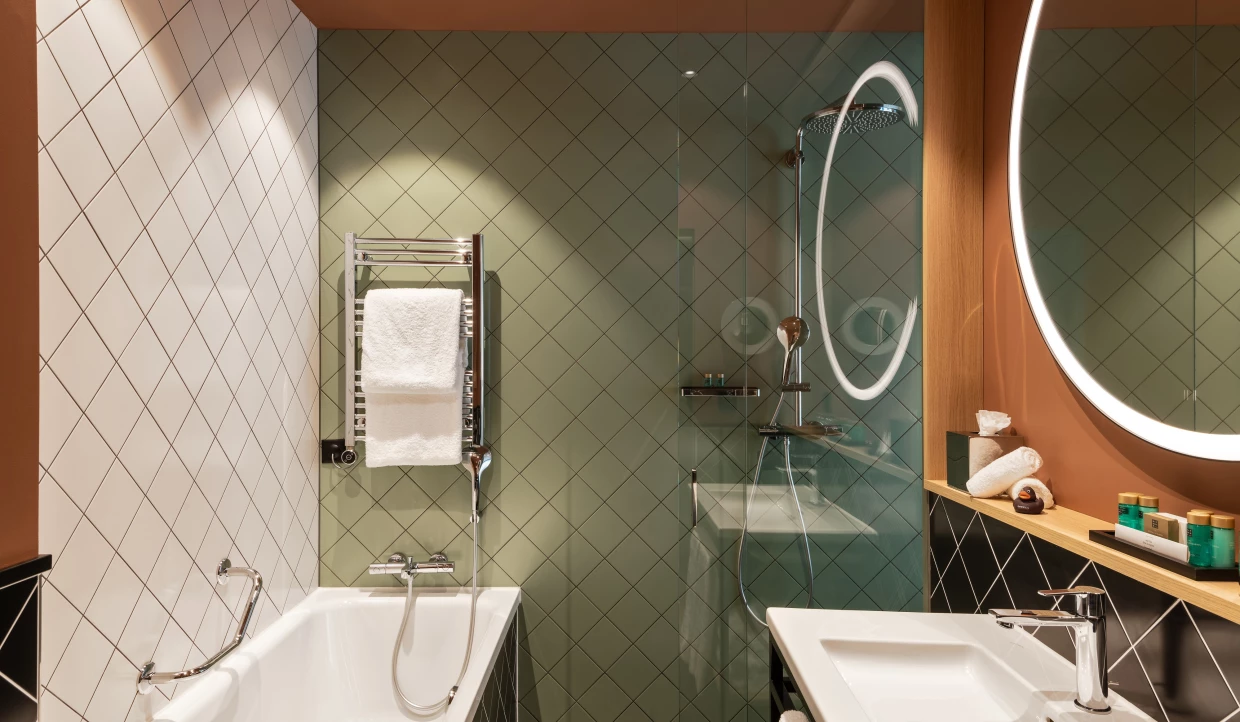 Badezimmer mit Badewanne, diagonalen Fliesen in grün, salbei und terracotta. Beleuchteter runder Spiegel.