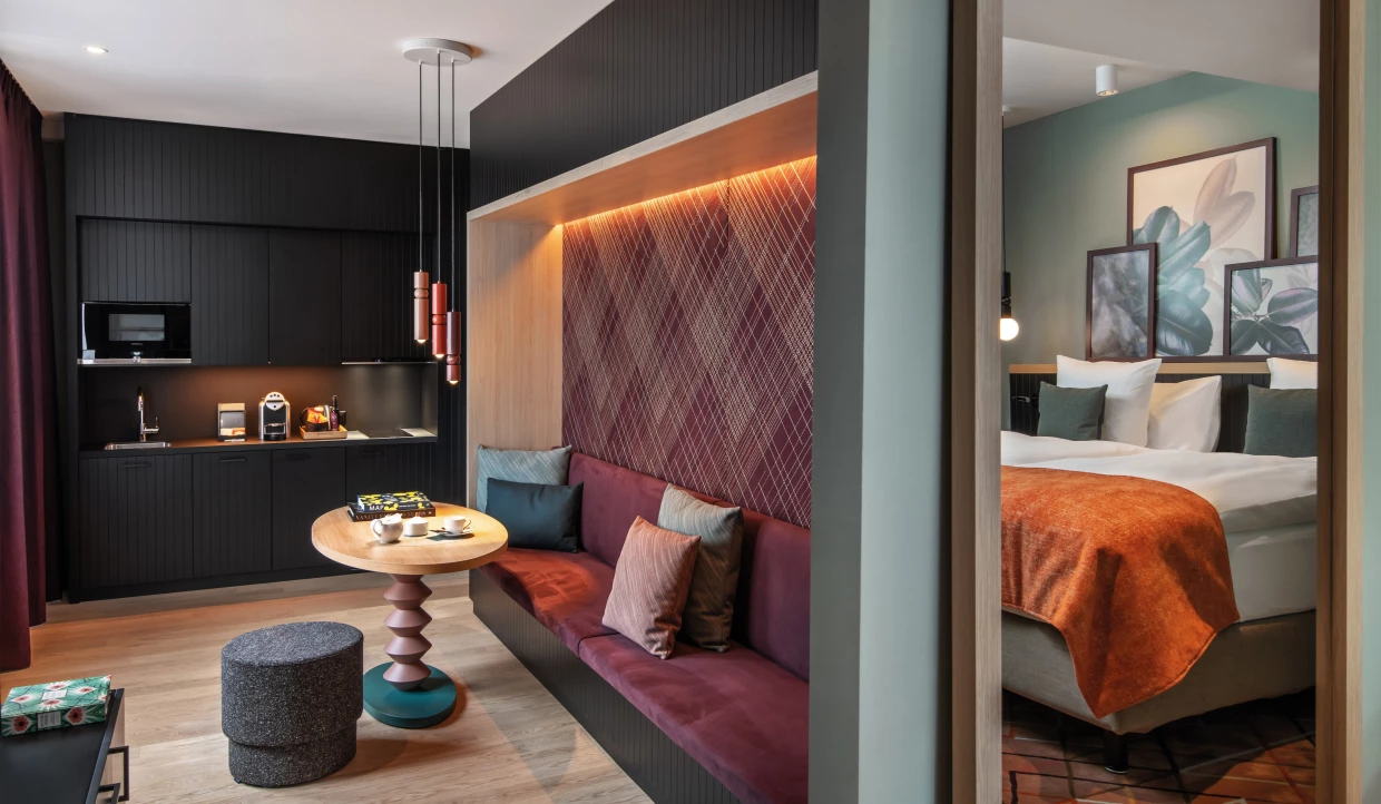 Design-Hotel-Apartment mit Küchenzeile und Sitznische links sowie Doppelbett mit Pflanzengrafiken am Betthaupt rechts.